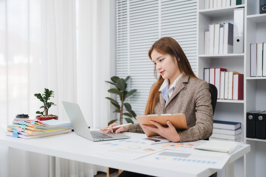 Focused professional at her desk: A young businesswoman attentively works on her laptop while holding a folder, surrounded by office supplies and neatly organized shelves.
