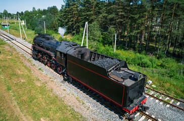 Aerial view captures an old black steam locomotive moving along parallel train tracks next to a dense green forest © Evgenii Bakhchev