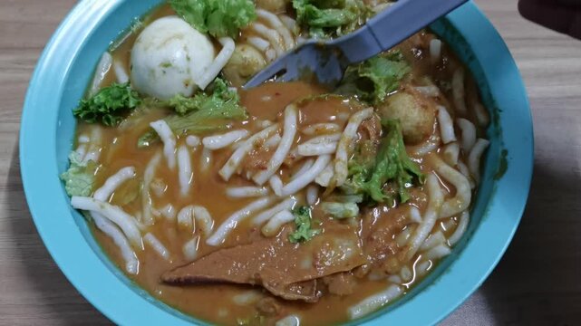 Spicy curry laksa noodles served in a light blue bowl with a fork, boiled egg, and lettuce greens