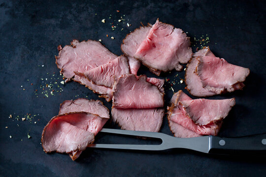 Six slices of roast beef, herbes and meat fork on dark ground