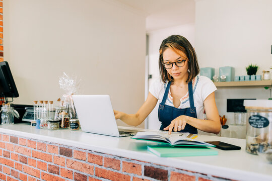Entrepreneur working on laptop and checking book at shop counter