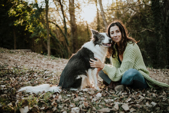 Smiling woman sitting in autumn forest petting her border collie dog