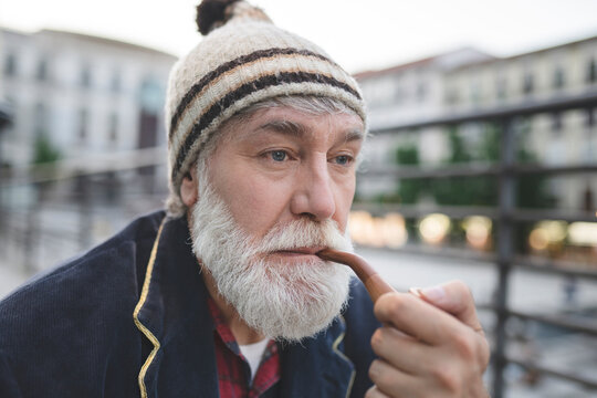 Thoughtful mature man with white beard wearing knit hat smoking pipe