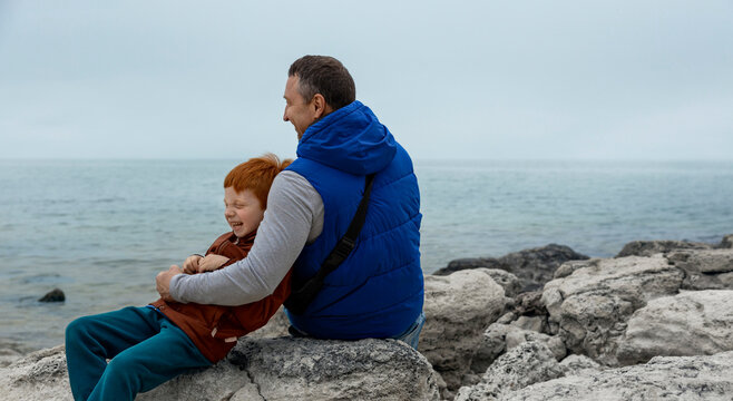 A happy father and son sit on stones by the sea
