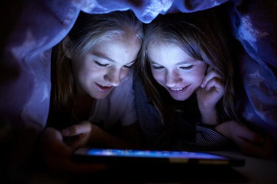 Two sisters playing secretly with their digital tablet, hidden under blanket