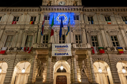 Torino, Italia - 13/12/2014 - piazza Palazzo di Citt&agrave;. Facciata storica illuminata con banner multilingue e simboli europei. Scena urbana notturna ideale per temi accoglienza e architettura.