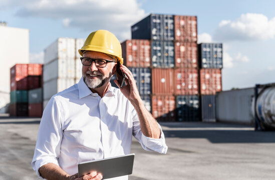 Businessman at cargo harbour, wearing safety helmet, using smartphone and digital tablet