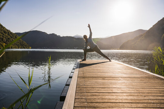 Woman exercising with hand raised on jetty over lake by mountains