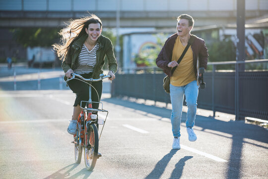 A smiling woman rides a bicycle while a man runs in the city