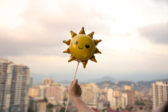 Hand of woman holding sun shaped smiley balloon against skyand city skyline