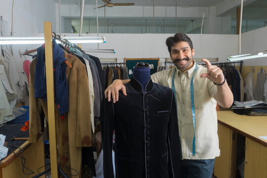 Tailor standing beside a mannequin wearing a semi-stitched coat in his workshop and showing a gesture of holding a product in fingers.