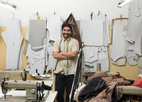 Smiling tailor standing with arms crossed in his small workshop with sample cuttings hanging in the background.