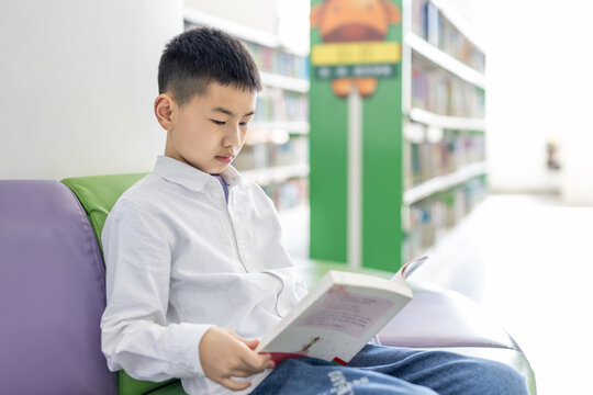 A young Asian boy sits on a chair in a library, focusing on reading an open book.