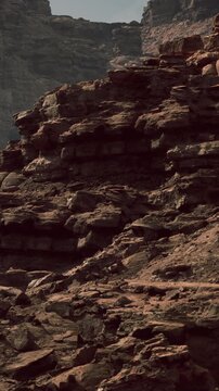 Rocks and stones create a rough surface in a dry landscape under a clear sky. Sunlight highlights the layers of the rock formation and the texture in the ground.