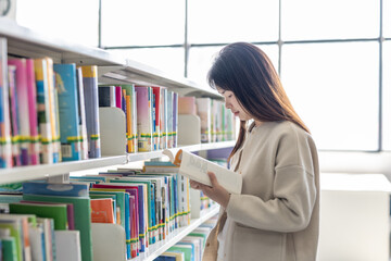 A young woman with long hair reads a book while standing by bookshelves in a bright library. © tai