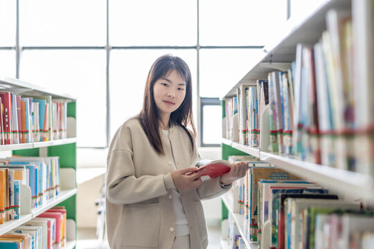 An east asian young woman stands between library bookshelves, holding and reading a red book in a bright indoor space.