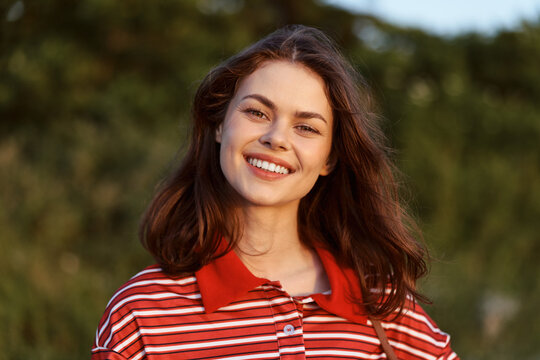 Smiling brunette woman posing outdoors during sunset. Lifestyle portrait showing a happy female model with natural makeup and striped clothing. Soft lighting creates a warm summer atmosphere.