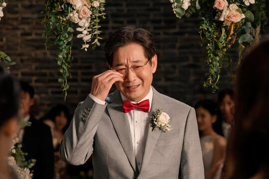 A tearful middle-aged Asian man in a gray suit cries during an indoor wedding ceremony, surrounded by floral decor and guests.
