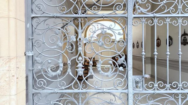 A slow upward camera movement follows an ornate wrought iron entrance gate, revealing intricate scrollwork, handcrafted metal details. Slow tilt-up shot filmed from close range.