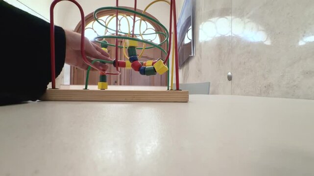 A close-up shows a hand gently guiding colorful wooden beads along curved metal rails on a classic wire maze activity. Close-up static shot filmed from the front at table height.