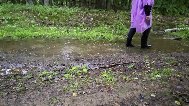Person walking through muddy puddle in forest environment, wearing black boots and purple raincoat, splashing water during a wet day outdoors, demonstrating resilience and exploration.