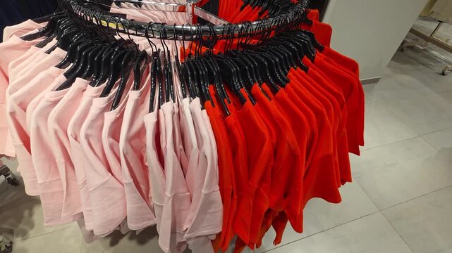 Person hand browsing selection of pink t-shirts on circular clothing rack displaying variety of colorful garments including vibrant red and soft pink shirts in retail clothing store environment