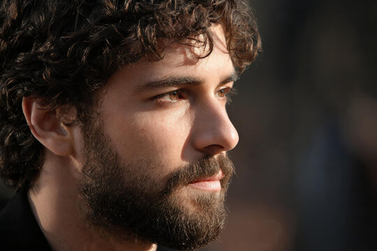 A close-up side portrait of an adult man with curly dark hair and a full beard, captured in warm natural lighting.