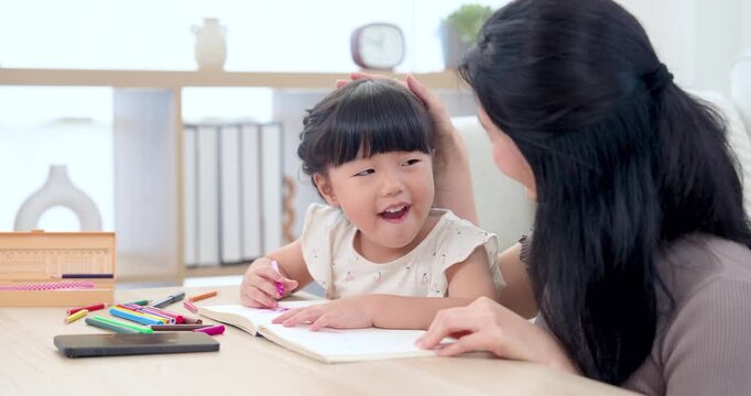 Asian single mother gently guiding preschool daughter drawing in workbook at home, smiling child holding pink pencil during early learning play, warm family bonding, nurturing care, creative growth
