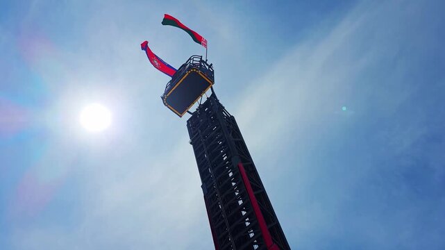 Fire truck ladder extended high into clear blue sky sun flare, rescue platform two colorful flags waving from top, low angle perspective emergency equipment reaching sky