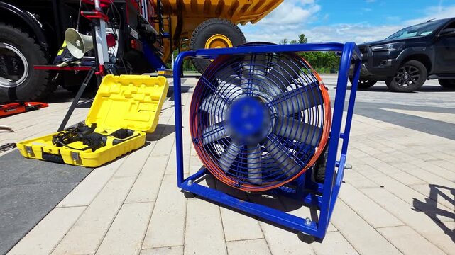 Large blue industrial fan with orange trim and yellow toolbox with tools on paved ground next to heavy machinery and a vehicle under a clear sky on a bright day.