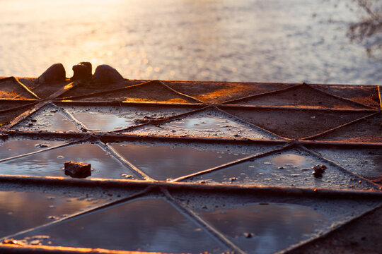 Close-up of a weathered metal pontoon dock with geometric steel frames and puddles, capturing the soft warm glow of a setting sun reflecting off the tranquil river surface at twilight