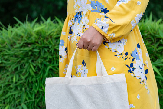 Woman Holding Blank Cotton Tote Bag in Yellow Floral Dress Against Green Foliage