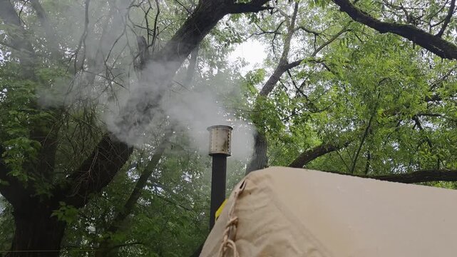 Smoke rising from metal chimney of canvas tent in lush green forest during overcast day outdoor camping adventure temporary shelter for wilderness exploration