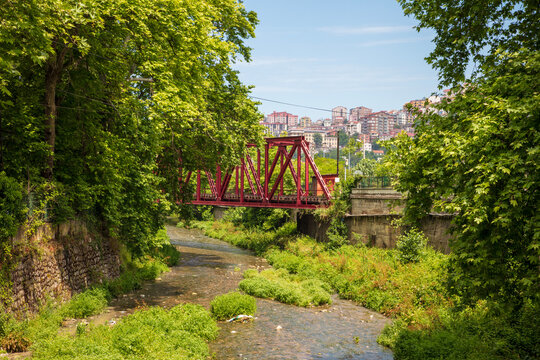 Historic red steel railway bridge spanning a small stream in Zonguldak, Turkey, surrounded by lush green trees with the city's hillside skyline in the background.