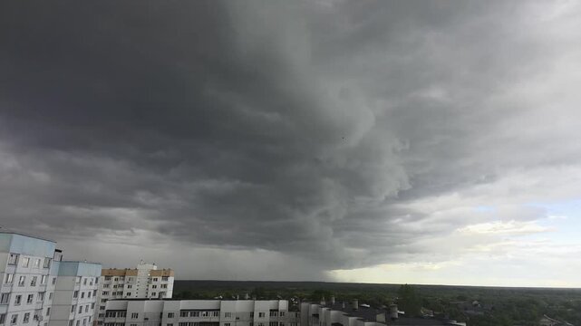 Dark storm clouds gather over city buildings and distant green forest, promising heavy rain and dramatic weather changes, creating a moody atmosphere.