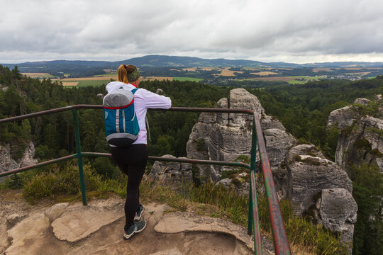 Woman with a backpack stands at a railing on a rock lookout in Bohemian Paradise, Czech Republic, looking at the forest