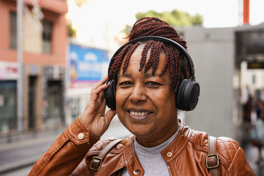 Happy senior african woman using wireless headphones while listening music in the city - Elderly person, technology and travel concept