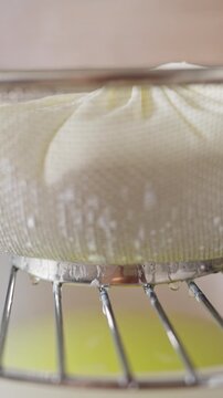 Vertical video. Homemade Cottage Cheese Draining Through Cheesecloth in a Metal Strainer Above a Bowl in a Kitchen. Traditional Dairy Making and Healthy Nutrition