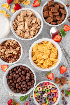 Variations of breakfast cereals in bowls. Bowl with corn pads, colorful rings, chocolate balls and cornflakes on a gray concrete background. Top view, copy space.