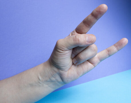 Close-up of a human hand making the &ldquo;rock on&rdquo; or &ldquo;horns&rdquo; gesture against a purple and blue background, symbolizing energy, music, enthusiasm, and freedom of expression.