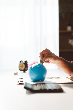 Hand placing coin into a blue piggy bank on a desk, representing concepts of saving money for the future, financial planning, budget management, and investment