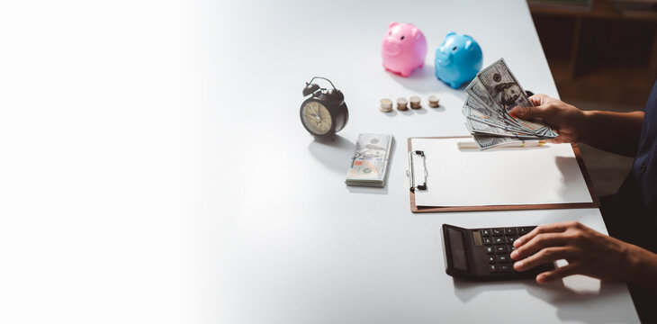 Person calculating budget and managing personal finances, holding us dollar cash with piggy banks, coins, and an alarm clock representing savings and time investment