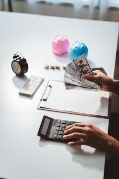 Person's hands counting dollar banknotes with a calculator, managing finances for savings and investment, with piggy banks and coins on a white desk for budgeting