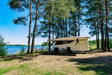 Camping trailer parked under tall pine trees near a serene blue lake on a sunny day © NBLX