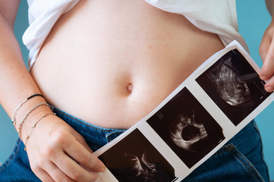 pregnant woman holding three ultrasound photos against belly in blue jeans closeup, concept of family, motherhood, prenatal care