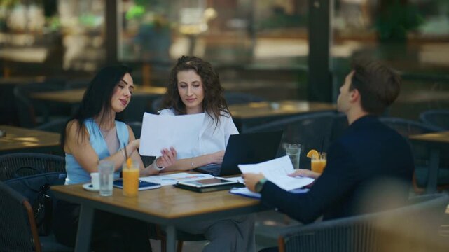 Business professionals engage in an important meeting at an outdoor cafe setting. They are seen collaborating and reviewing documents on a sunny day.