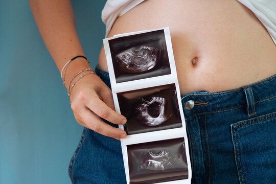 pregnant woman holding three ultrasound photos against belly in blue jeans closeup, concept of family, motherhood, prenatal care