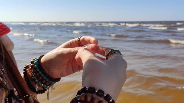 Hands wearing multiple rings hold a small white shell beside a sandy shoreline with rolling waves. 