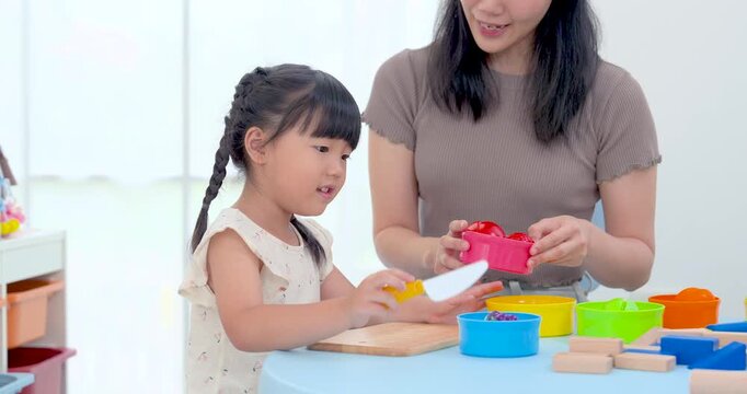 Asian mother hands toy pan toward preschool daughter during pretend kitchen game at table, encouraging role play, color matching, fine motor practice, conversation, and creative development at home.