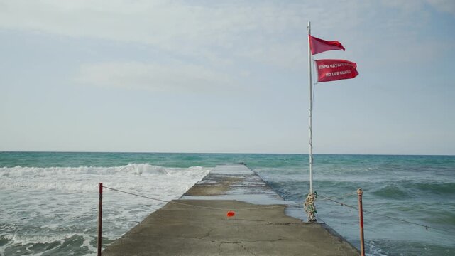 A red warning flag flutters in the wind on a wet concrete pier extending into the rough ocean, signaling dangerous waters.